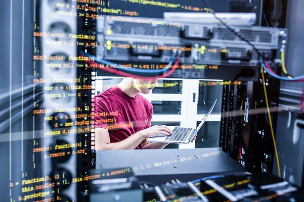 A person is working on a laptop in a server room, surrounded by server racks and network technology.