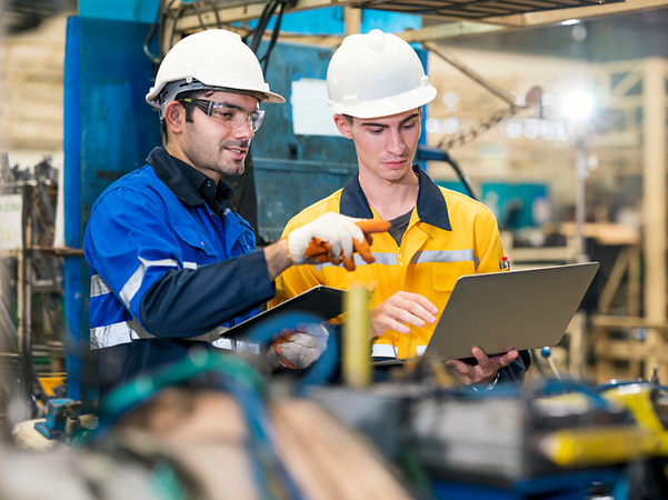  Zwei Arbeiter mit Schutzhelmen prüfen Daten auf einem Laptop in einer Fabrik