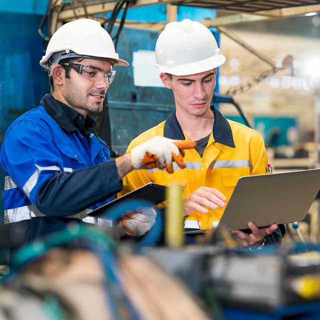  Zwei Arbeiter mit Schutzhelmen prüfen Daten auf einem Laptop in einer Fabrik