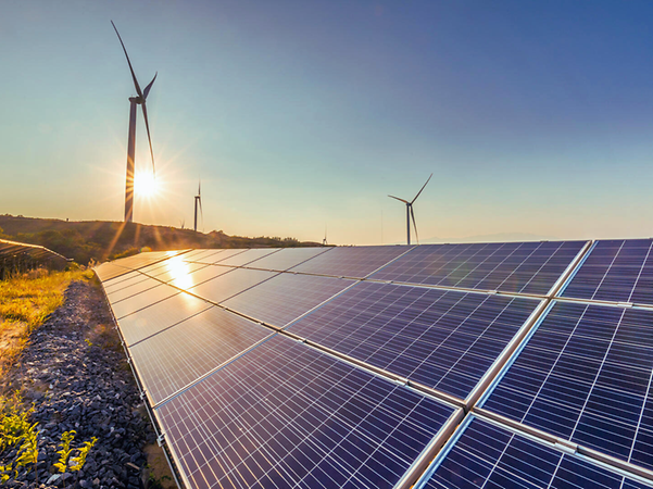 Solar modules in a field reflecting the sun, with wind turbines in the background.