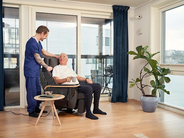 Pflegekraft steht neben einer älteren Person, die auf einem Stuhl sitzt.