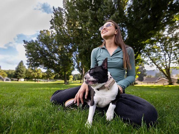Frau sitzt im Gras und streichelt ihren Hund, der ein Halsband trägt.