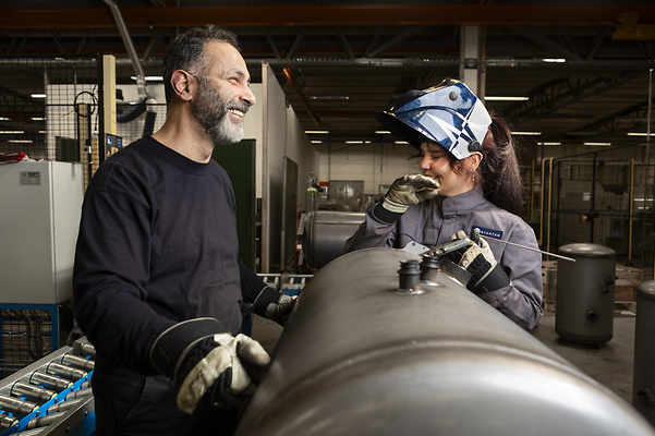 Zwei Mitarbeitende in einer Produktionshalle arbeiten gemeinsam an einem Metallbauteil und tauschen sich lächelnd aus.