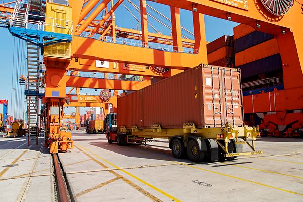 Ein Containerlager mit mehreren orangefarbenen Portalkränen und einem LKW, der einen langen Container transportiert.