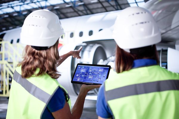 Two women wearing safety gear and holding a tablet point at an airplane inside a hangar